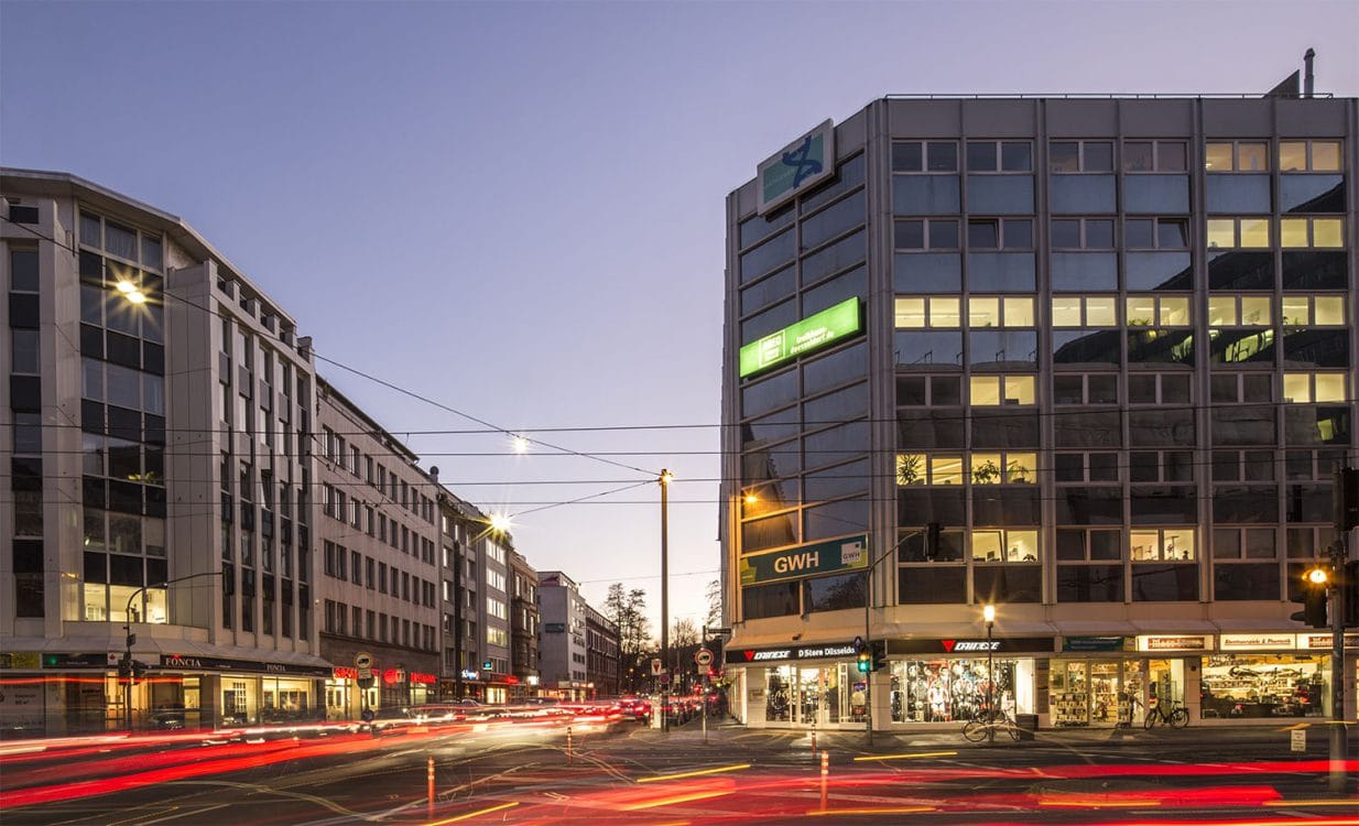 Straßenkreuzung in der Abenddämmerung, links ein helles Geschäftshaus, rechts ein verglastes Eckgebäude, davor Lichtspuren des Verkehrs.