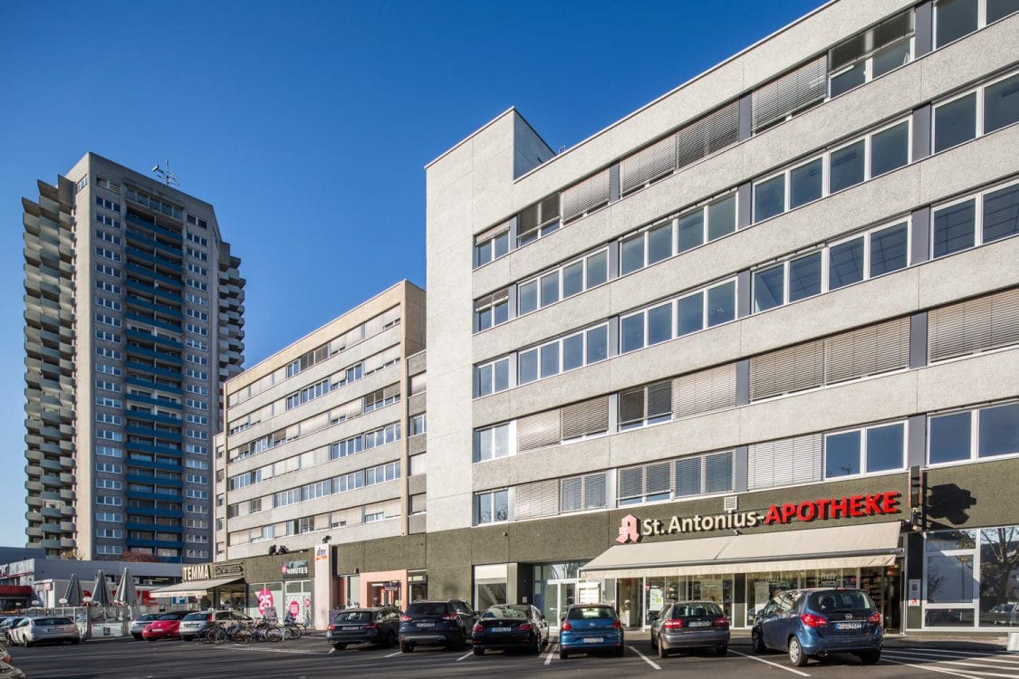 Langgestrecktes mehrgeschossiges Gebäude mit Apotheke und Ladenzeile im Erdgeschoss, davor ein Parkplatz, links ein Wohnhochhaus.