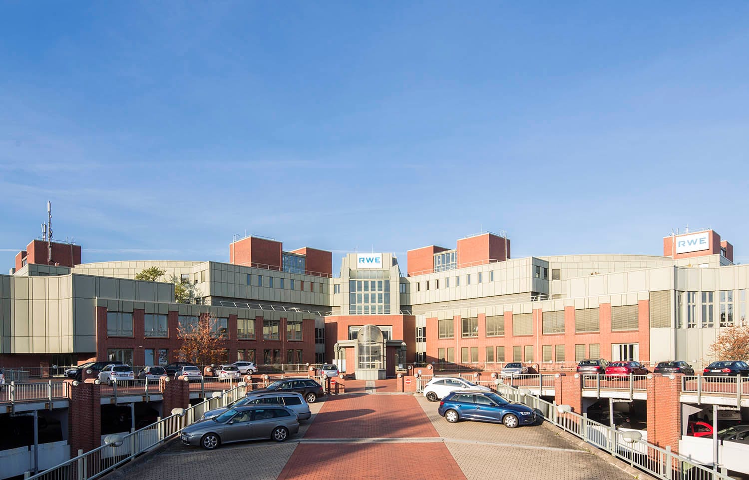 Weitläufiger Bürokomplex aus Backstein und hellen Metallpaneelen mit vorgelagerten Parkdecks und gepflasterter Zufahrt.