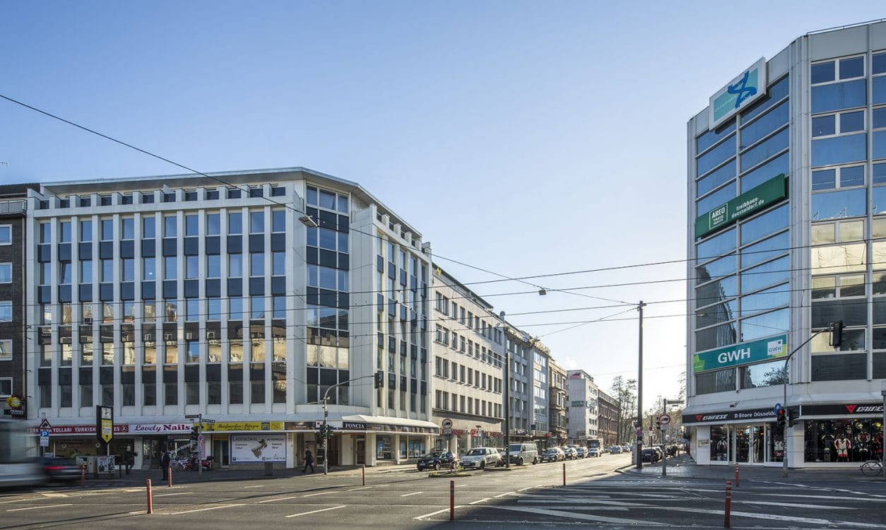 Innerstädtische Kreuzung mit einem hellen Geschäftshaus links und einem verglasten Eckgebäude rechts, dazwischen Straßenbahnoberleitungen.