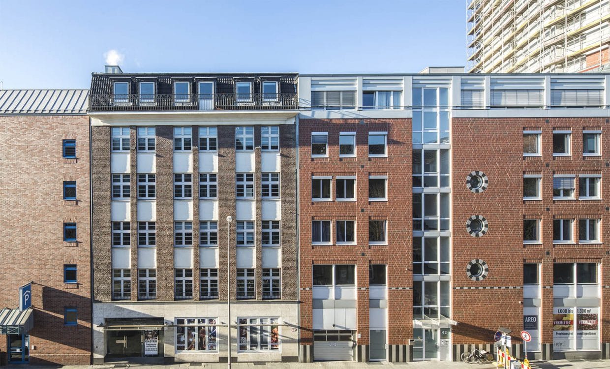Straßenzeile aus Backsteinhäusern mit hellen Fensterachsen und dunklem Staffelgeschoss, im Hintergrund ein eingerüstetes Hochhaus.