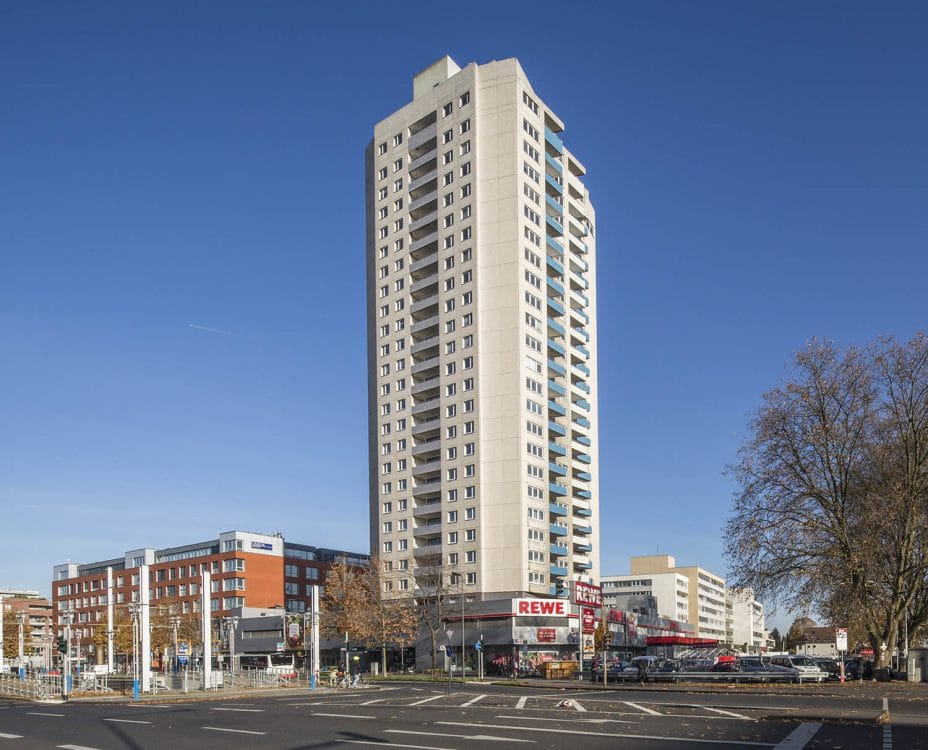 Schlankes Wohnhochhaus mit vorspringenden Balkonen über einer eingeschossigen Ladenzeile, davor eine breite Straßenkreuzung.