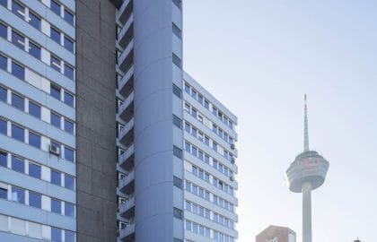 Hochformatige Ansicht eines Bürohochhauses mit hellblauer Rasterfassade und rundem Treppenturm, im Hintergrund ein Fernsehturm.