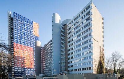 Bürogebäude mit hellblauer Rasterfassade und runden Treppentürmen, links ein Hochhaus mit farbig gemustertem Fassadenmosaik.