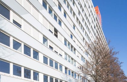 Fluchtperspektive entlang der langen Fassade eines Bürogebäudes mit umlaufenden Fensterbändern, davor Rasenstreifen und kahle Bäume.