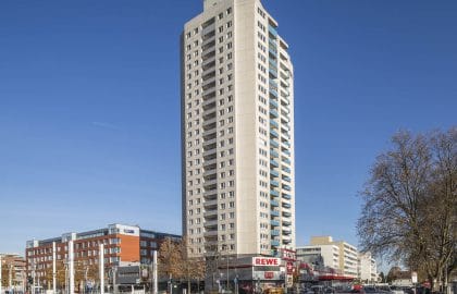 Schlankes Wohnhochhaus mit vorspringenden Balkonen über einer eingeschossigen Ladenzeile, davor eine breite Straßenkreuzung.