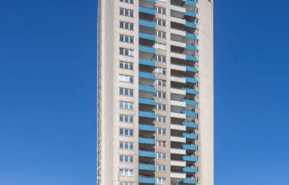 Untersicht auf ein vielgeschossiges Wohnhochhaus mit blau verkleideten Balkonbrüstungen über einer Supermarktzeile.