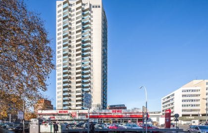 Wohnhochhaus mit gestaffelter Betonfassade über einem breiten Supermarktbau, links herbstlich gefärbtes Laub, davor ein Parkplatz.