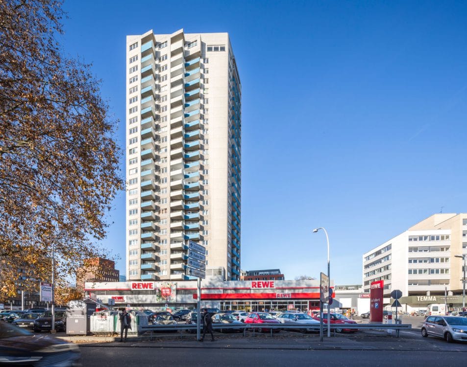 Wohnhochhaus mit gestaffelter Betonfassade über einem breiten Supermarktbau, links herbstlich gefärbtes Laub, davor ein Parkplatz.