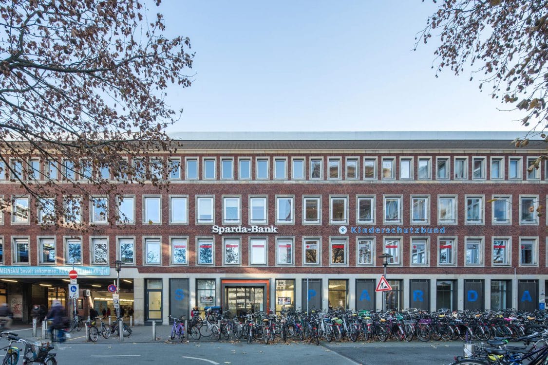 Viergeschossiges Backsteingebäude mit Bankfiliale im Erdgeschoss, davor Hunderte abgestellter Fahrräder und herbstliches Laub.