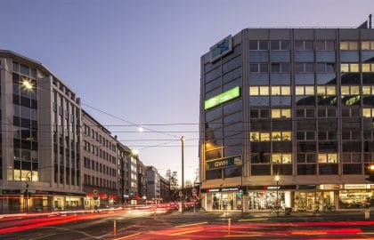 Straßenkreuzung in der Abenddämmerung mit einem verglasten Eckgebäude, Ladenlokalen im Erdgeschoss und Lichtspuren des Verkehrs.