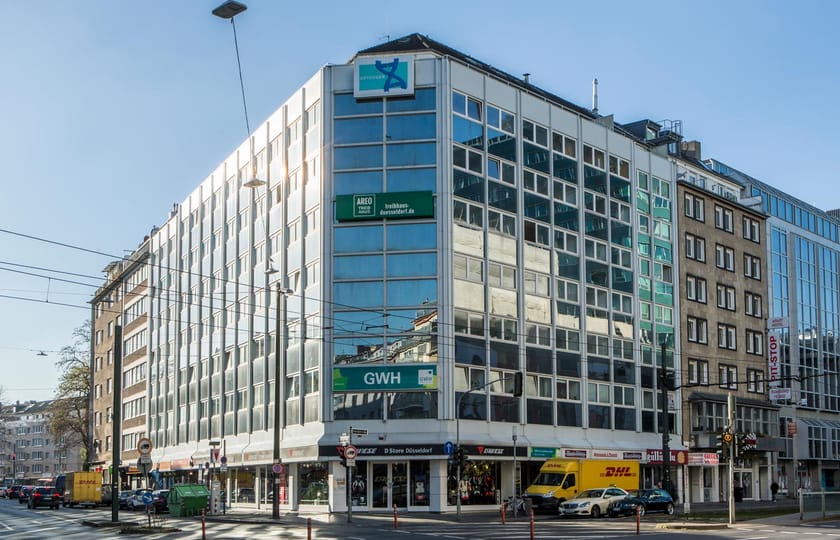 Verglastes Eckgebäude mit weißer Rasterfassade über einer Ladenzeile, an einer Kreuzung mit Straßenbahnoberleitungen bei Sonnenschein.