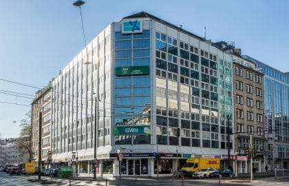Verglastes Eckgebäude mit weißer Rasterfassade über einer Ladenzeile, an einer Kreuzung mit Straßenbahnoberleitungen bei Sonnenschein.