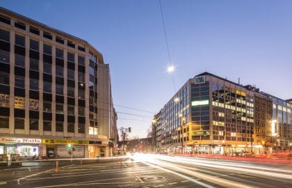 Weite Kreuzung in der blauen Stunde, links ein helles Geschäftshaus, rechts ein beleuchtetes Eckgebäude, dazwischen Lichtspuren.