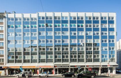 Frontale Ansicht einer weißen Rasterfassade über einer durchgehenden Ladenzeile, davor parkende Autos bei klarem Himmel.