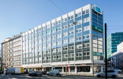 Schräge Straßenansicht eines Geschäftshauses mit weißer Rasterfassade, davor fließender Verkehr in Bewegungsunschärfe.