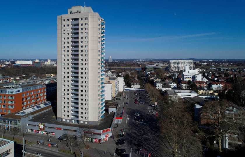 Luftaufnahme eines hellen Wohnhochhauses mit Balkonen über einer Ladenzeile, ringsum niedrige Stadtbebauung bis zum Horizont.