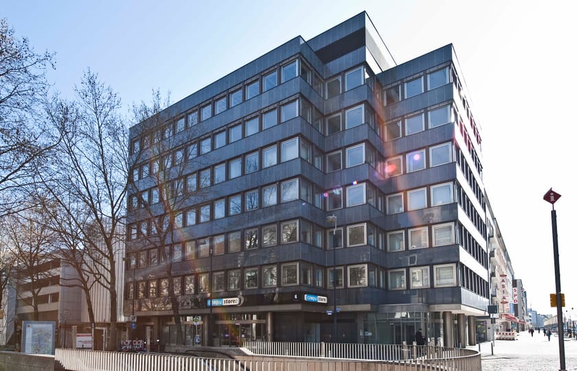 Mehrgeschossiges Eckgebäude mit dunkler Fassadenverkleidung und Ladenlokalen im Erdgeschoss, an einem innerstädtischen Platz.