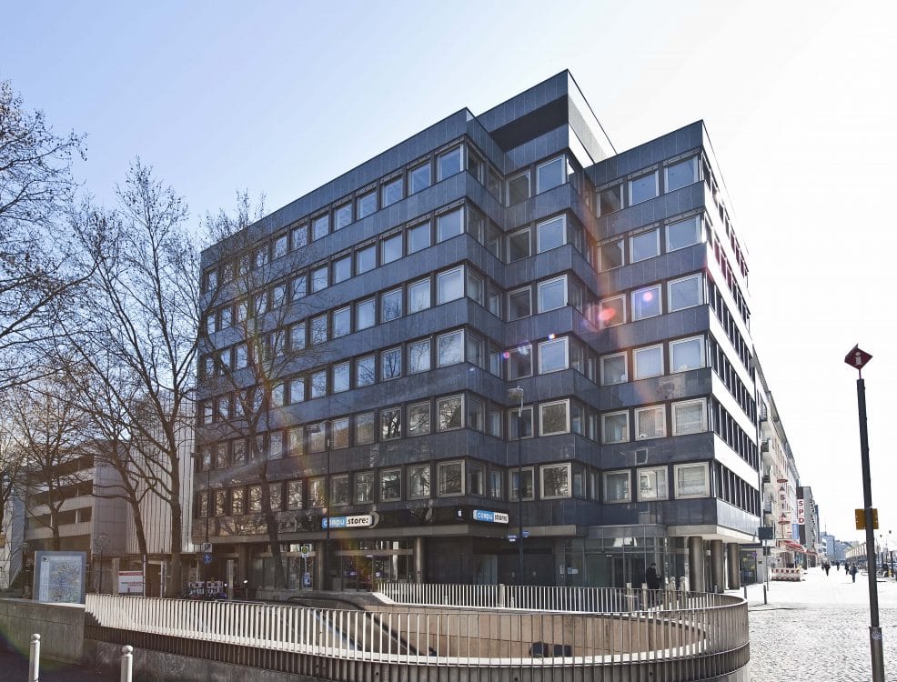Mehrgeschossiges Eckgebäude mit dunkler Fassadenverkleidung und Ladenlokalen im Erdgeschoss, an einem innerstädtischen Platz.