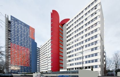 Rückseite eines Bürokomplexes mit weißen Fensterbändern und rotem, rundem Treppenturm, links ein Hochhaus mit farbigem Fassadenmosaik.