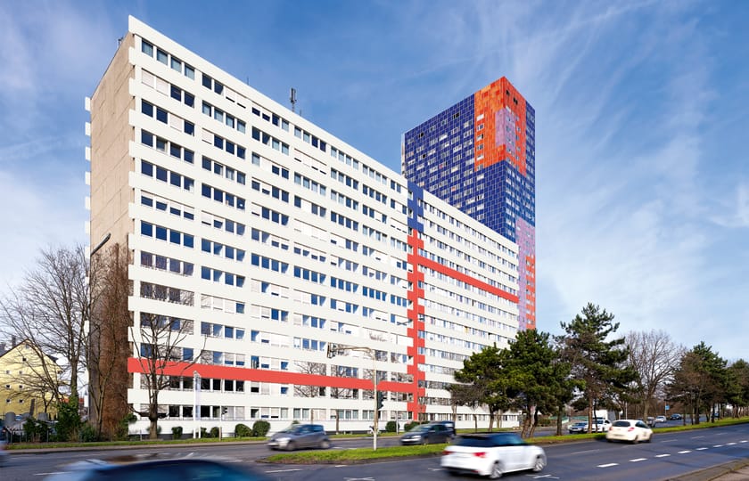 Langgestrecktes weißes Bürogebäude mit roten Fassadenbändern an einer befahrenen Straße, dahinter ein blau-orange gemustertes Hochhaus.