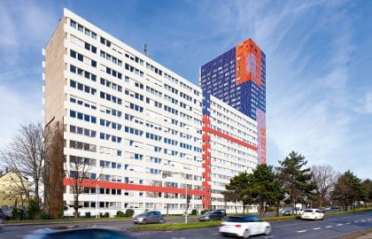 Long white office building with red facade bands along a busy road, a blue-and-orange patterned high-rise rising behind it.