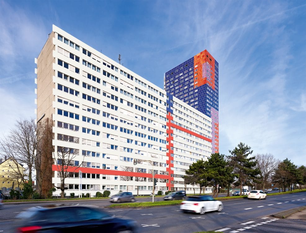 Langgestrecktes weißes Bürogebäude mit roten Fassadenbändern an einer befahrenen Straße, dahinter ein blau-orange gemustertes Hochhaus.