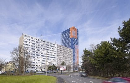 Blick über eine geschwungene Straße auf einen weißen Gebäuderiegel und ein dahinter aufragendes Hochhaus mit blau-oranger Fassade.