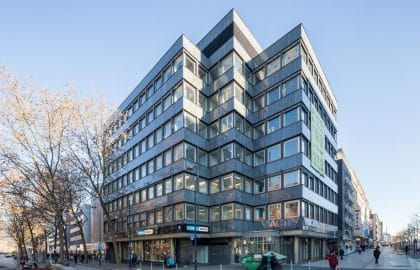 Eckgebäude mit dunkel verkleideter Fassade und breiten Bandfenstern, im Erdgeschoss Ladenlokale an einer Fußgängerstraße.