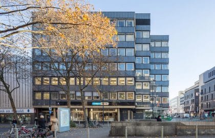Frontale Ansicht eines dunkel verkleideten Geschäftshauses hinter einem herbstlich gefärbten Baum, davor ein gepflasterter Platz.