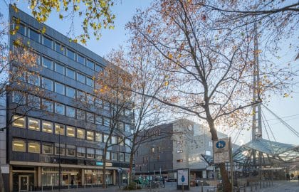 Dunkel verkleidetes Bürogebäude hinter herbstlichem Baumbestand, rechts anschließend ein Bau mit gläsernem Vordach.