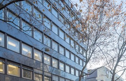 Fluchtansicht entlang der dunklen Fassade eines Bürogebäudes mit Ladenzeile und Arkade im Erdgeschoss.