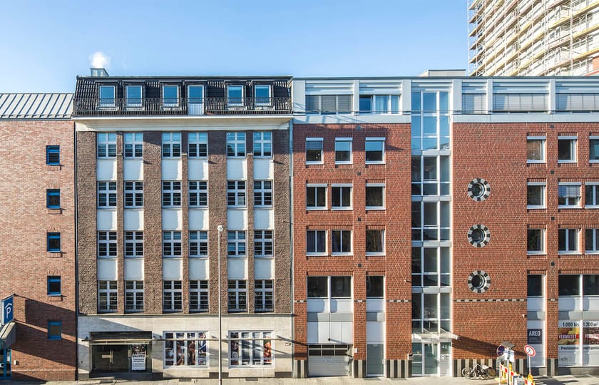 Straßenzeile aus Backsteinhäusern mit hellen Fensterachsen und dunklem Staffelgeschoss, im Hintergrund ein eingerüstetes Hochhaus.