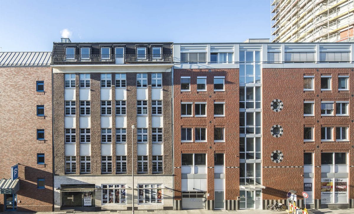 Straßenzeile aus Backsteinhäusern mit hellen Fensterachsen und dunklem Staffelgeschoss, im Hintergrund ein eingerüstetes Hochhaus.