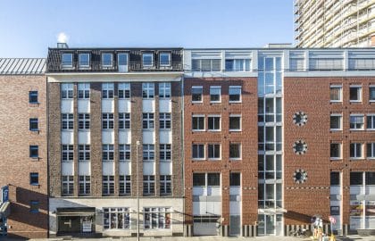 Straßenzeile aus Backsteinhäusern mit hellen Fensterachsen und dunklem Staffelgeschoss, im Hintergrund ein eingerüstetes Hochhaus.