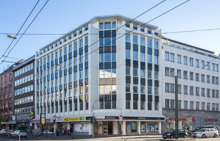 Weißes Eckgebäude mit senkrechten Fassadenrippen und Ladenlokalen im Erdgeschoss, an einer Kreuzung mit Straßenbahnoberleitungen.