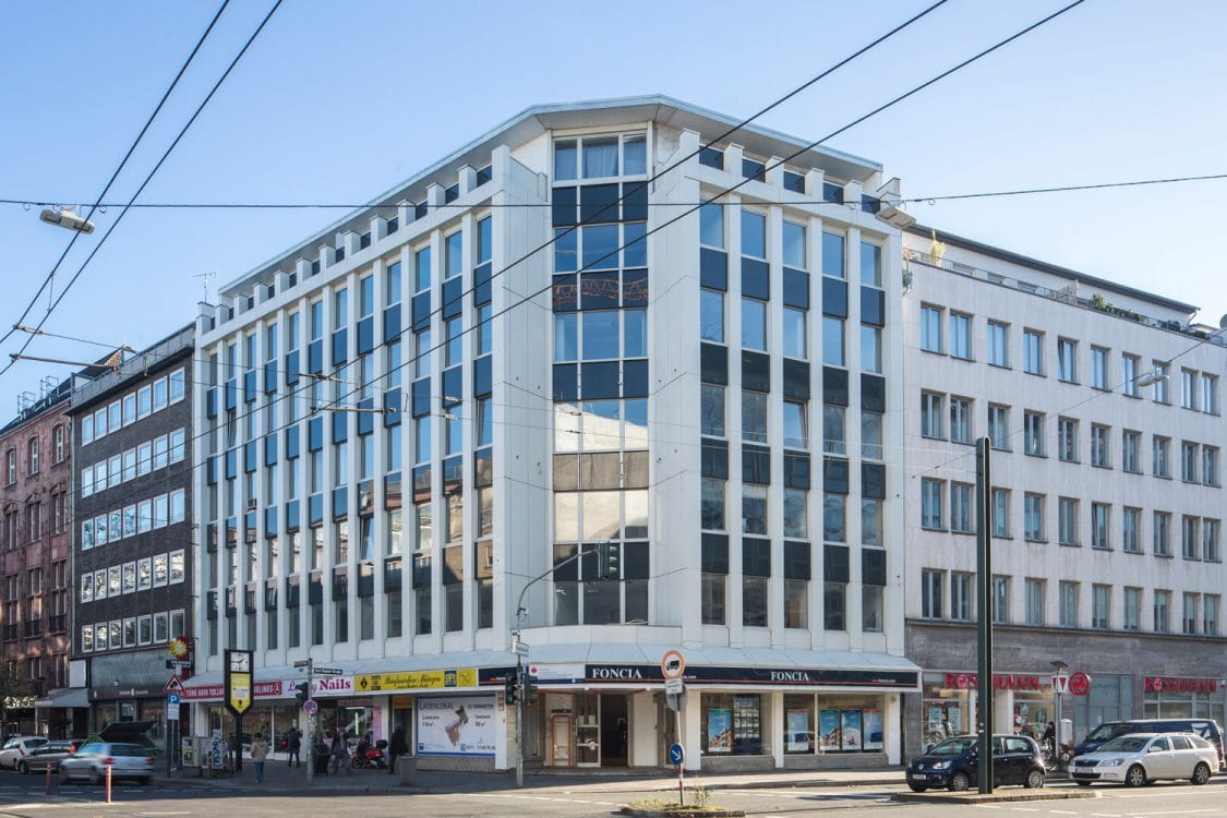 Weißes Eckgebäude mit senkrechten Fassadenrippen und Ladenlokalen im Erdgeschoss, an einer Kreuzung mit Straßenbahnoberleitungen.