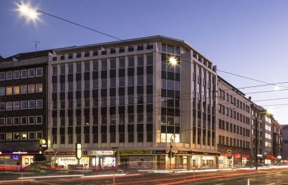 Weißes Eckgebäude mit Fassadenrippen in der Abenddämmerung, darunter eine beleuchtete Ladenzeile und Lichtspuren des Verkehrs.