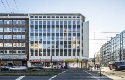 Frontale Straßenansicht eines hellen Geschäftshauses mit großflächiger Verglasung zwischen weißen Lisenen, links ein dunklerer Altbau.