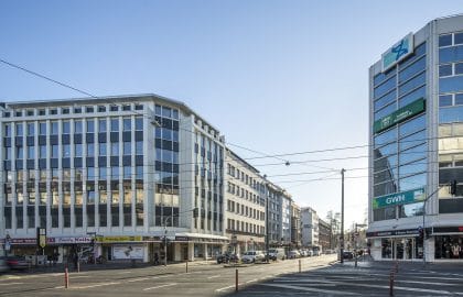 Innerstädtische Kreuzung mit einem hellen Geschäftshaus links und einem verglasten Eckgebäude rechts, dazwischen Straßenbahnoberleitungen.