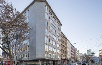 Mehrgeschossiges Eckhaus mit fensterloser Betongiebelwand und Ladenlokalen im Erdgeschoss, an einer Straße mit Straßenbahngleisen.