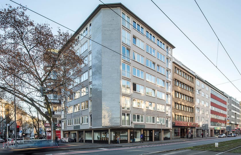 Eckgebäude mit heller Betonfassade und geschlossener Giebelwand, dahinter eine Häuserzeile entlang einer breiten Straße.