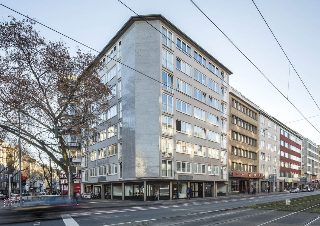 Eckgebäude mit heller Betonfassade und geschlossener Giebelwand, dahinter eine Häuserzeile entlang einer breiten Straße.