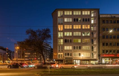Beleuchtetes mehrgeschossiges Gebäude in der blauen Stunde mit gewerblich genutztem Erdgeschoss, davor eine Kreuzung mit Lichtspuren.