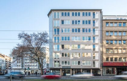 Frontale Ansicht eines hellen mehrgeschossigen Gebäudes mit regelmäßigen Fensterreihen, davor vorbeifahrender Verkehr in Bewegungsunschärfe.