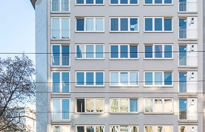 Narrow multi-storey building with a pale rendered facade, slim balconies and commercial units at street level.