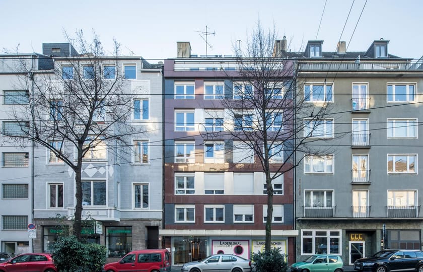 Straßenzeile aus drei aneinandergrenzenden Wohnhäusern, das mittlere mit rotbraun-grauer Plattenfassade und leerem Ladenlokal.