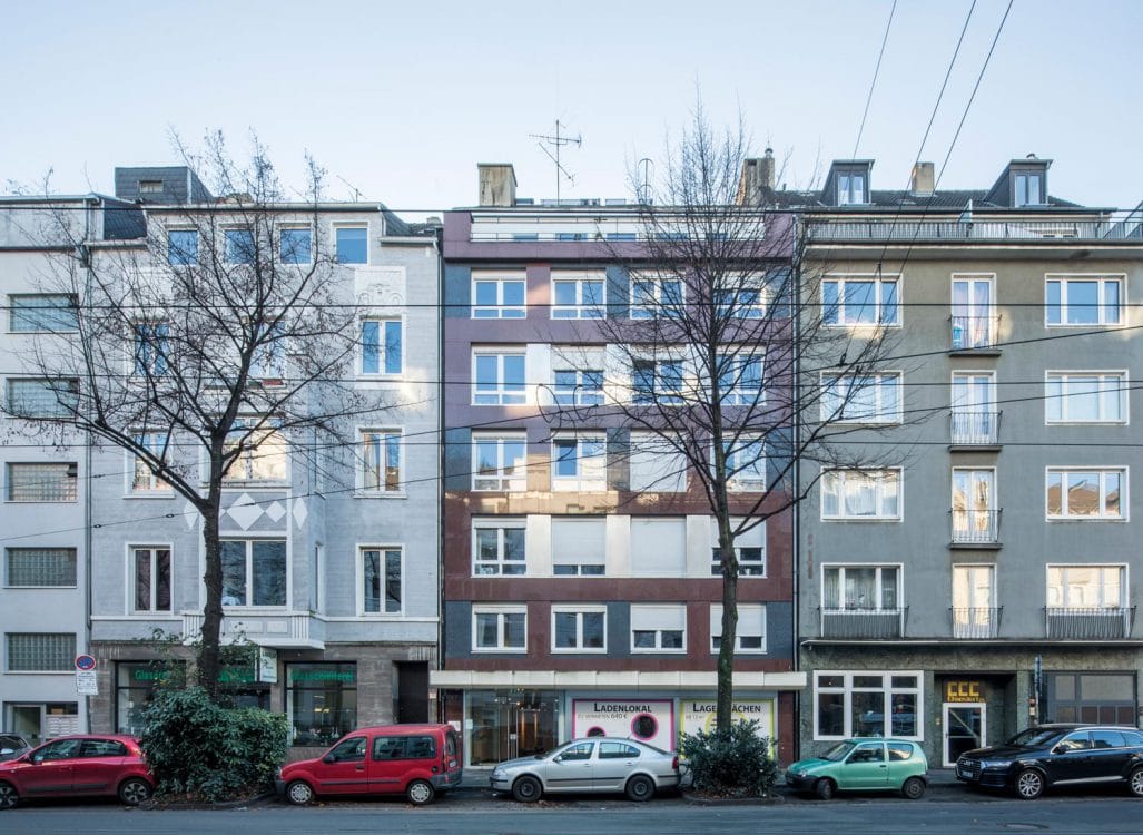 Straßenzeile aus drei aneinandergrenzenden Wohnhäusern, das mittlere mit rotbraun-grauer Plattenfassade und leerem Ladenlokal.