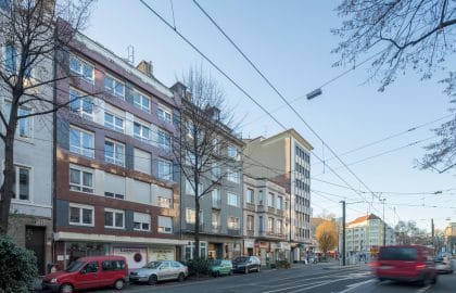 Schrägansicht einer Häuserzeile mit farbig verkleideter Fassade und Ladenlokalen, entlang einer Straße mit Oberleitungen.