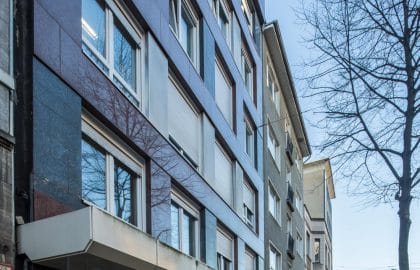 Hauseingang mit polierter Steinverkleidung und Vordach, daneben ein Ladenlokal mit Werbeplakaten an einer Wohnstraße.